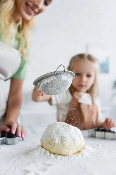 Niño borroso tamizar harina en masa cruda cerca de mamá sonriente en la cocina - foto de stock