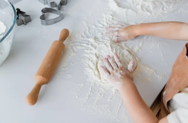 Vista de ángulo alto del niño recortado amasando masa cerca de rodillo y cortadores de galletas - foto de stock