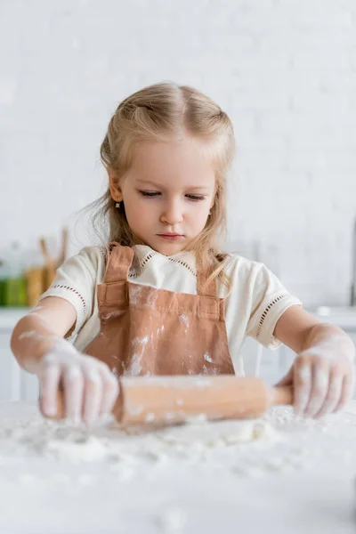 Kid in apron rolling dough in kitchen on blurred foreground — Stock Photo