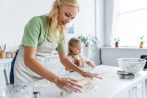 Frau mit kleiner Tochter rollt rohen Teig auf Küchentisch — Stockfoto