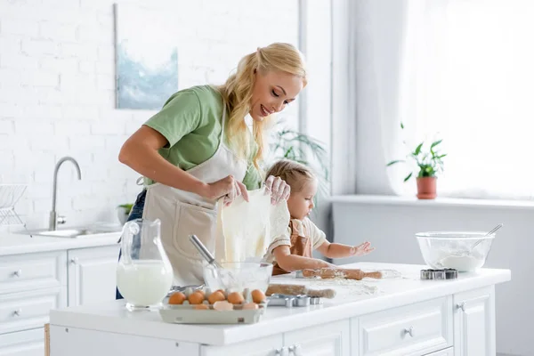 Mujer sonriente sosteniendo masa casera mientras cocina con su hija en la cocina - foto de stock