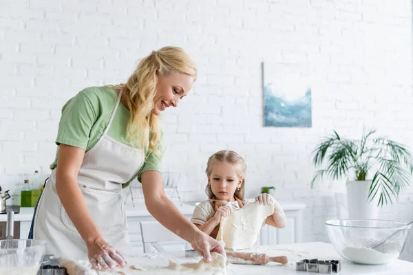 Маленькая девочка держит сырое тесто, помогая маме на кухне — стоковое фото