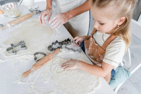 Vista de ángulo alto de la chica de corte de masa con cortador de galletas cerca de mamá en la cocina - foto de stock