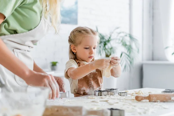 Petite fille tenant la pâte près de mère floue et emporte-pièce sur la table de cuisine — Photo de stock