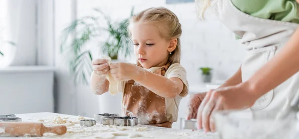 Girl in apron near blurred mother cutting raw dough with cookie cutter, banner — Stock Photo