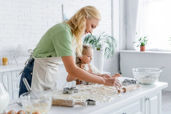 Femme avec fille cuisinière dans la cuisine près de la table avec pâte crue et ustensiles de cuisine — Photo de stock