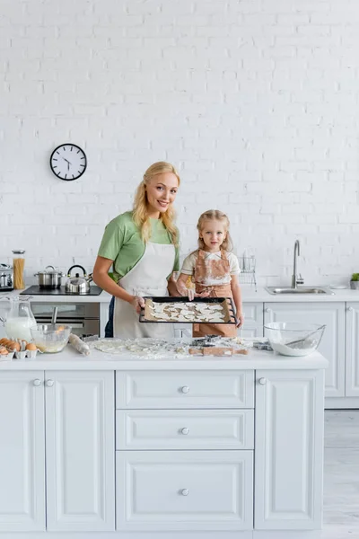 Mulher sorridente com filha olhando para a câmera perto da assadeira com biscoitos crus — Fotografia de Stock