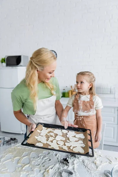 Maman et fille souriant à l'autre près de plaque à pâtisserie avec des biscuits crus — Photo de stock