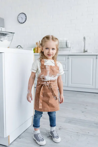 Chica con manchas de harina en el delantal de pie en la cocina - foto de stock