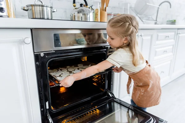 Chica en delantal colocación de bandeja para hornear con galletas en el horno - foto de stock
