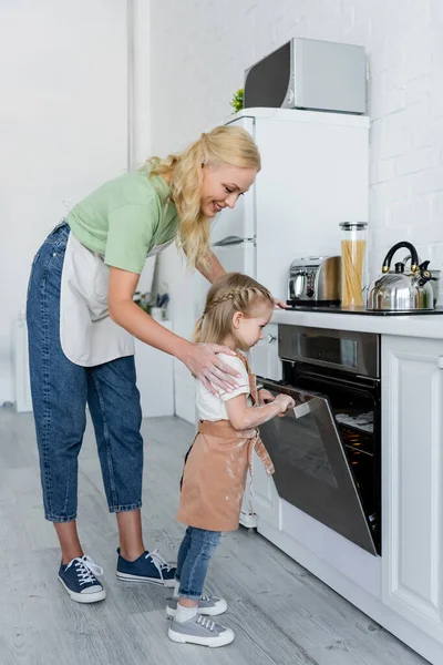 Mädchen sieht Kekse im Elektroofen neben glücklicher Mutter — Stockfoto