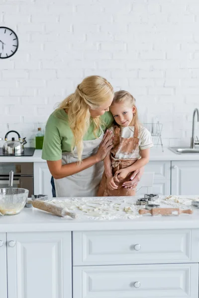 Mujer abrazando hija cerca de la mesa de la cocina con harina y utensilios de cocina - foto de stock