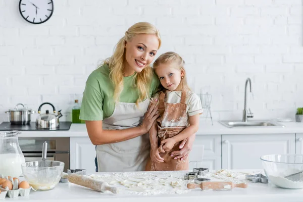 Mujer feliz abrazo niño cerca de la mesa de la cocina con ingredientes y utensilios de cocina - foto de stock