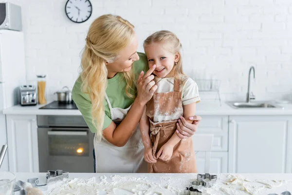 Woman touching nose of cheerful daughter near kitchen table with dough and cookie cutters — Stock Photo