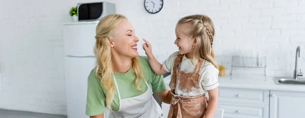 Chica en delantal con manchas de harina tocando la nariz de mamá feliz en la cocina, pancarta - foto de stock