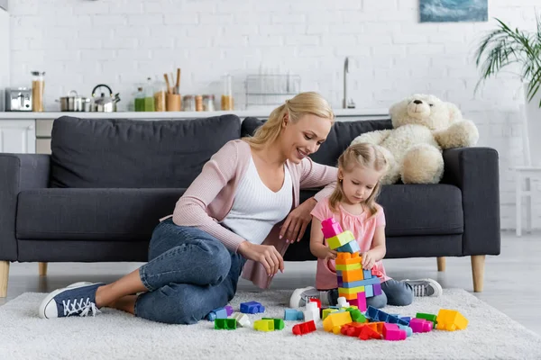 Mädchen bastelt Turm aus Bauklötzen in der Nähe von Mama und Teddybär auf Couch — Stock Photo