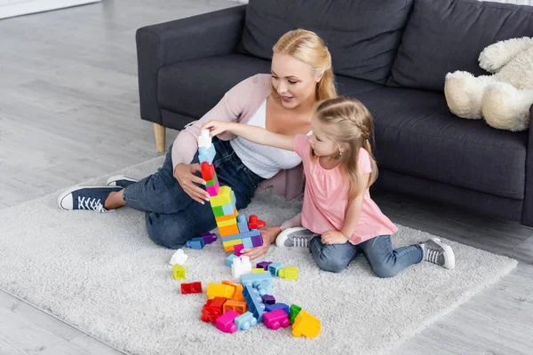 Niño haciendo torre de bloques de construcción cerca de mamá en el piso en casa - foto de stock