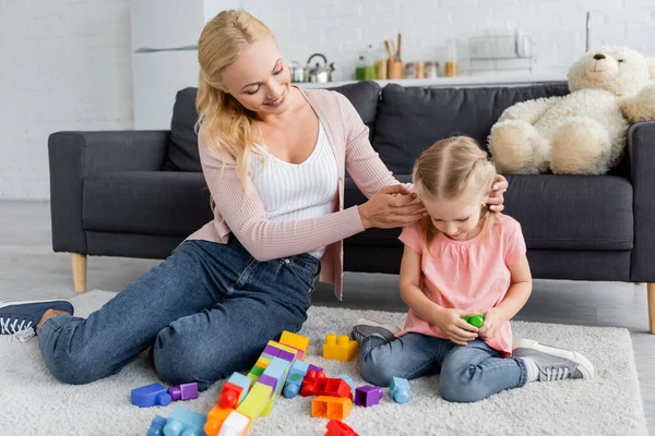 Lächelnde Frau justiert Haare ihrer Tochter, die zu Hause mit Bauklötzen auf dem Boden spielt — Stockfoto