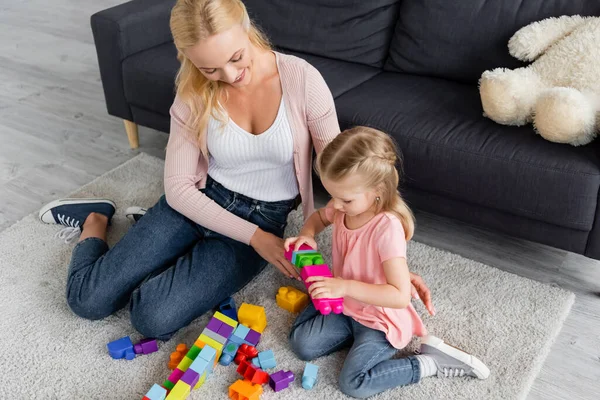 Vista de ángulo alto de la niña jugando con bloques de construcción de colores en el piso cerca de la madre - foto de stock