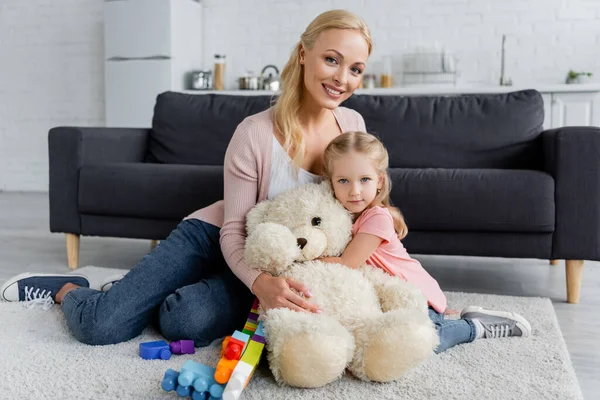 Mother and daughter embracing on floor near teddy bear and colorful building blocks — Stock Photo