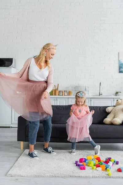 Mulher feliz e menina em saias de tule e coroas de brinquedo dançando na cozinha — Fotografia de Stock