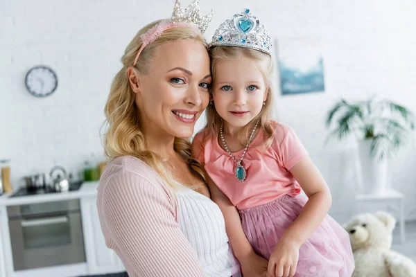 Feliz mamá e hija en coronas de juguete sonriendo a la cámara en casa - foto de stock