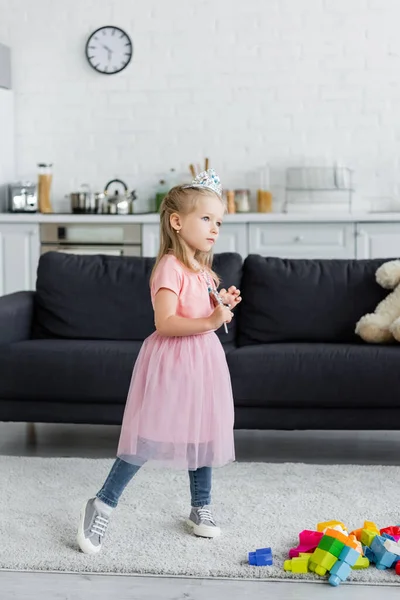 Menina em coroa de brinquedo e saia de tule de pé com varinha mágica em casa — Fotografia de Stock