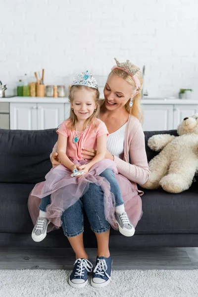 Cheerful mother and daughter in toy crowns sitting on sofa near teddy bear — Stock Photo