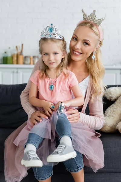 Mom and daughter in toy crowns smiling at camera while sitting on couch — Stock Photo