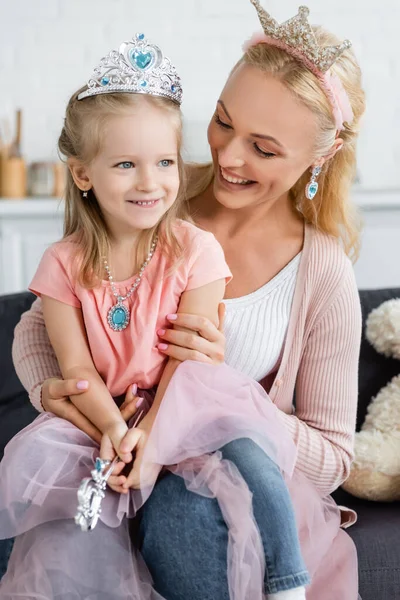 Cheerful mom and daughter in toy crowns, earrings and necklace sitting on couch at home — Stock Photo