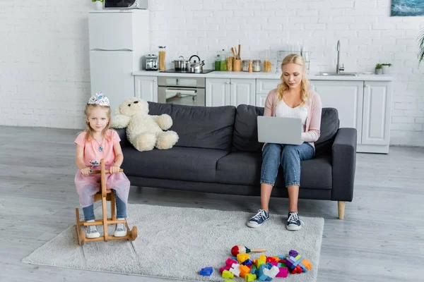 Menina em traje de princesa montando cavalo de balanço perto da mãe trabalhando no laptop no sofá — Fotografia de Stock