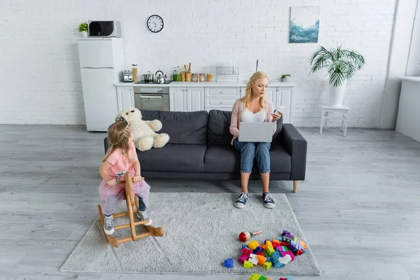 Mädchen reitet Schaukelpferd im Kostüm einer Prinzessin in der Nähe von Mama arbeitet auf Laptop auf Sofa — Stockfoto