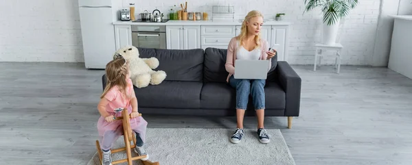 Frau mit Laptop und Smartphone neben Tochter auf Schaukelpferd — Stockfoto