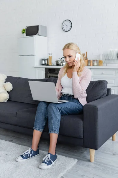 Blonde woman talking on mobile phone while sitting on sofa with laptop — Stock Photo