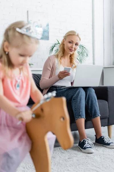 Woman with laptop and mobile phone looking at blurred daughter riding rocking horse — Stock Photo