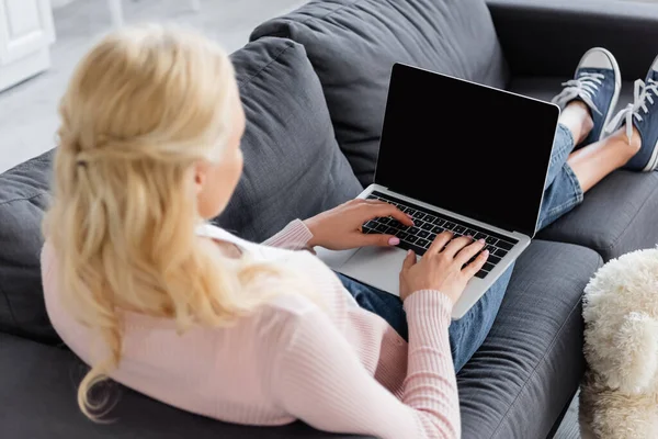Блондинка, печатающая на ноутбуке с пустым экраном, сидя на диване — стоковое фото