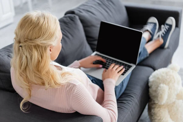 Блондинка сидить на дивані і друкує на ноутбуці з порожнім екраном — стокове фото