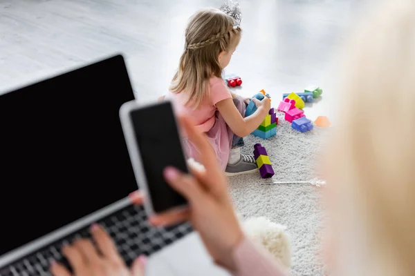 Niño jugando en el piso con bloques de construcción cerca de mamá con gadgets en primer plano borroso - foto de stock