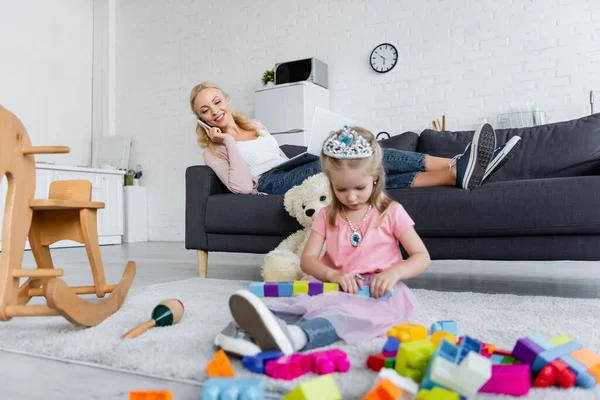 Smiling woman talking on smartphone near daughter in toy crown playing on floor — Stock Photo