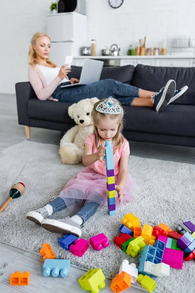 Chica en juguete corona jugando en el suelo con bloques de construcción cerca de mamá con gadgets sobre fondo borroso - foto de stock