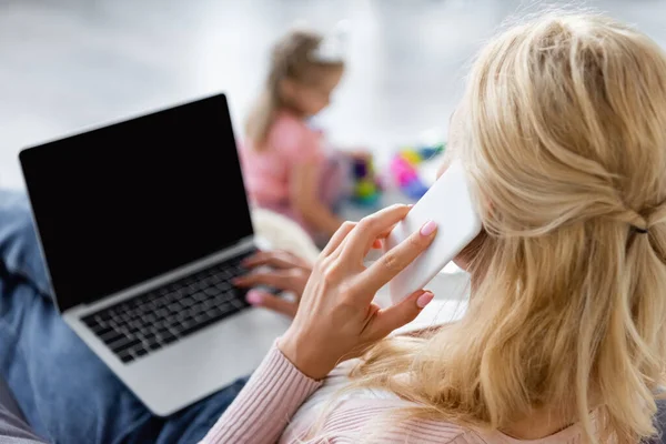 Mujer hablando en el teléfono inteligente cerca de la computadora portátil con la pantalla en blanco y la hija jugando sobre fondo borroso - foto de stock