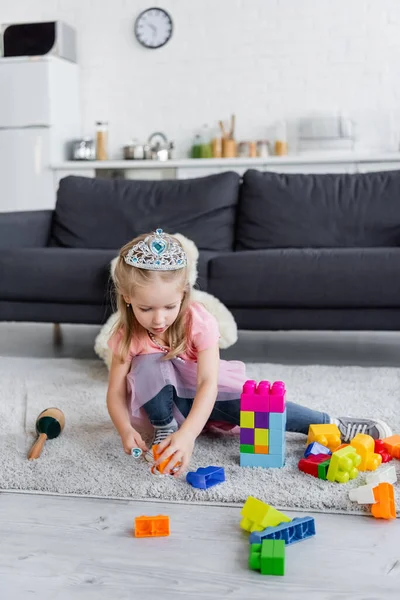 Kid in toy crown playing with multicolored building blocks on floor at home — Stock Photo