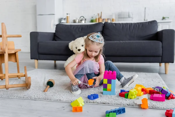 Chica en juguete corona jugando con varita mágica y cubos de colores en el suelo cerca de sofá y osito de peluche - foto de stock
