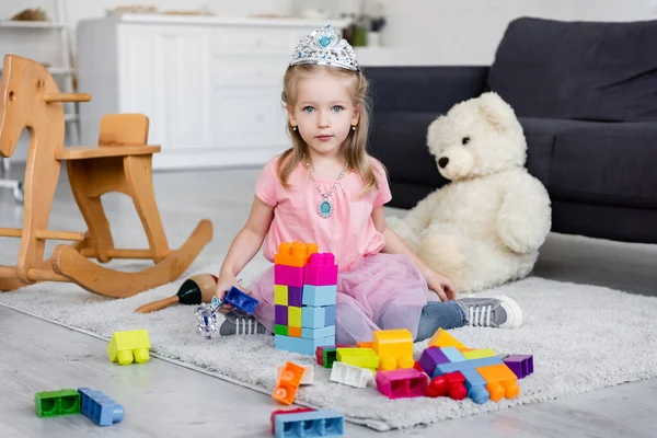 Criança em coroa de brinquedo e colar olhando para a câmera perto de blocos de construção e ursinho de pelúcia borrado — Fotografia de Stock