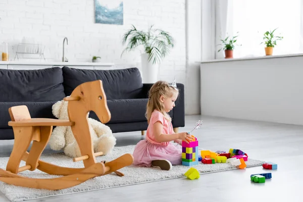 Girl in princess costume playing with magic wand and building blocks near rocking horse — Stock Photo