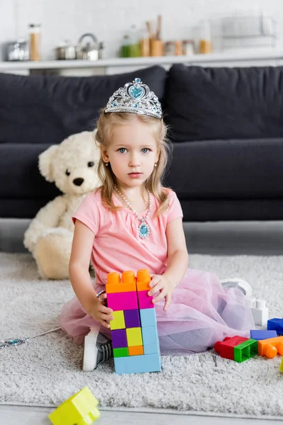 Girl in toy crown looking at camera near tower of colorful building blocks — Stock Photo