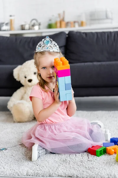 Menina em coroa de brinquedo mostrando torre de blocos de construção multicoloridos enquanto sentado no chão — Fotografia de Stock