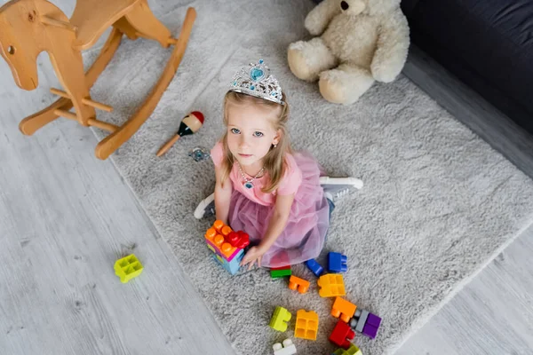 Overhead view of kid in toy crown playing with colorful building blocks near toys on floor — Stock Photo