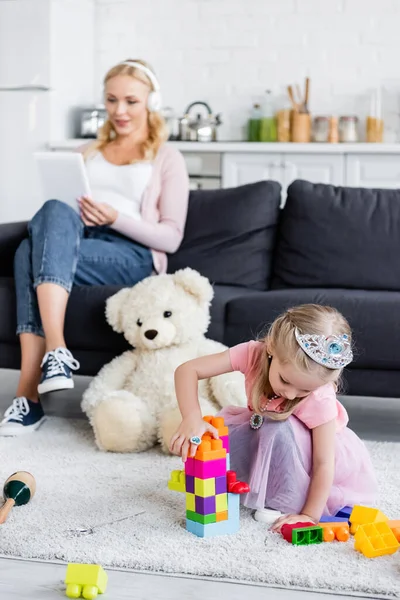 Fille dans jouet couronne faisant tour de blocs de construction tandis que maman écoute de la musique sur canapé — Photo de stock