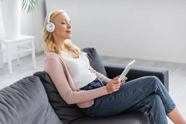 Donna bionda con tablet digitale ascoltare musica mentre seduta sul divano con gli occhi chiusi — Foto stock